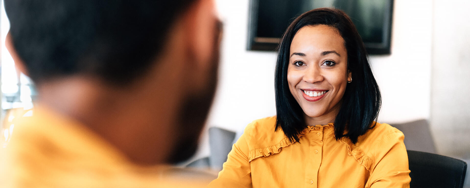 woman with tanned skin and straight hair, wearing a yellow long sleeve button down, smiling